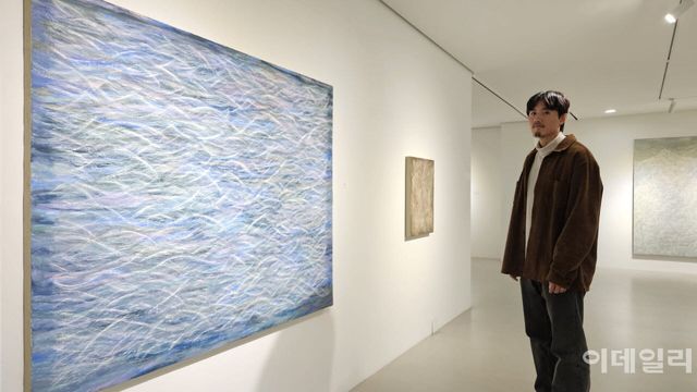 작가 우병윤이 선화랑 개인전 ‘중첩: 들떠 있는 풍경’에 건 자신의 작품들 앞에 섰다. 작가 정면에 ‘중첩 26-01-301’(2026, 162.0×130.3㎝)이 걸렸다. 무수하게 긁어낸 선과 선이 실타래처럼 엮였다. 나무패널 위의 석고가 드러나도록 날카로운 도구로 긁어낸 ‘상처’들이다(사진=오현주 문화전문기자).