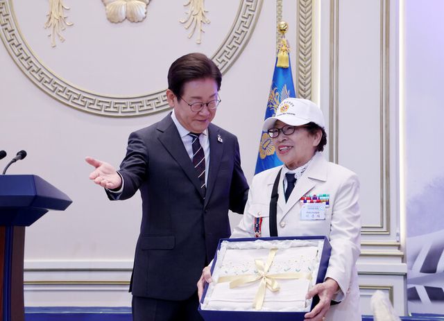 이재명 대통령이 27일 청와대 영빈관에서 열린 '호국 보훈의 달, 대통령의 초대' 행사에서 특별 초청대상자인 이춘자 씨에게 광목이불을 선물하고 있다. [사진=연합뉴스]