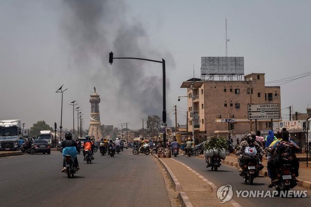 말리 수도 바마코의 건물에서 검은 연기가 피어오르는 가운데 오토바이가 거리를 지나고 있다.[AFP=연합뉴스. 재판매 및 DB 금지]