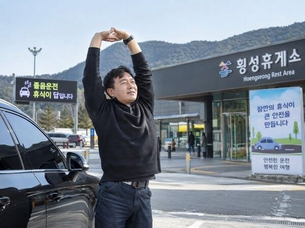 고속도로 휴게소에 차를 세운 운전자가 차량 밖으로 나와 기지개를 켜며 몸을 풀고 있다. 장거리 운전 중 졸음이 느껴질 때는 가까운 휴게소나 졸음쉼터에서 충분히 쉬어가는 것이 안전운전에 도움이 된다. 사진=AI 제작 이미지