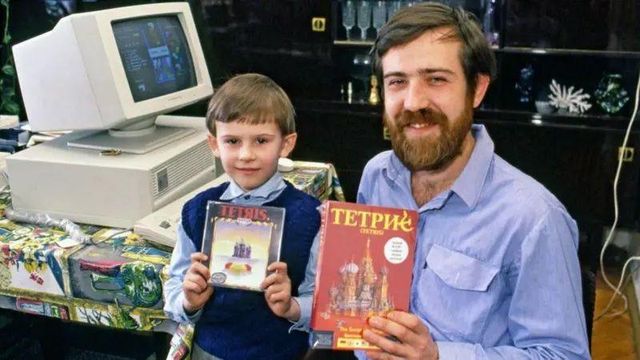 Alexey Pajitnov and a boy holding Tetris boxes. in front of a 1980s computer