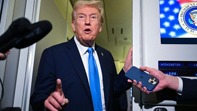 트럼프 대통령이 에어포스원 기내에서 기자들과 대화하고 있다. 그는 손가락으로 무언가를 가리키고, 옆에는 스크린이 보인다. 기자들은 녹음 장치를 들고 있다.