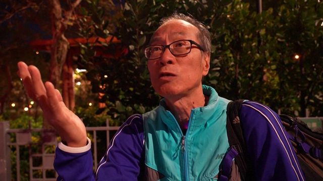 재킷을 입고 어깨에 배낭을 멘 남성이 카메라를 향해 말하고 있다