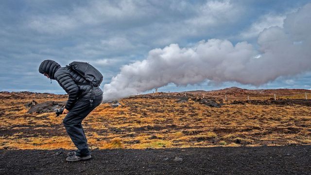 짧은 패딩 코트를 입고 후드를 쓴 남자가 워킹 팬츠와 신발을 신고 있는 모습을 담은 스톡 이미지. 그는 앞으로 쪼그려 앉아 아래쪽에서 구름이 터지는 것처럼 보이도록 자세를 취했다. 그는 풀밭 풍경 옆 도로에 있다