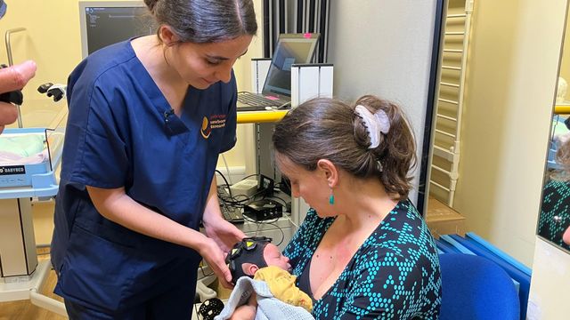 스타니 게오르기예바는 병원 내 영상 진단실의 의자에 앉아 아기 테오를 품에 안고 있다. 테오는 노란색 잠옷을 입고 파란색 담요에 싸여 있다. 플로라 포르 박사는 센서 캡을 막 장착한 뒤 테오를 향해 몸을 숙이고 있다.