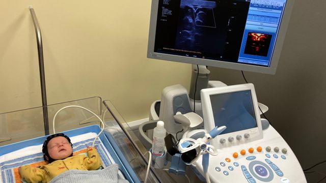 아기 테오는 병원용 아기 침대에 누워 잠든 상태다. 그의 머리에는 작은 검은색 센서 캡이 씌워져 있으며, 흰색 케이블을 통해 인근의 기계들과 연결되어 있다. 연결된 모니터에서는 테오의 뇌를 촬영한 영상이 실시간으로 표시되고 있다.