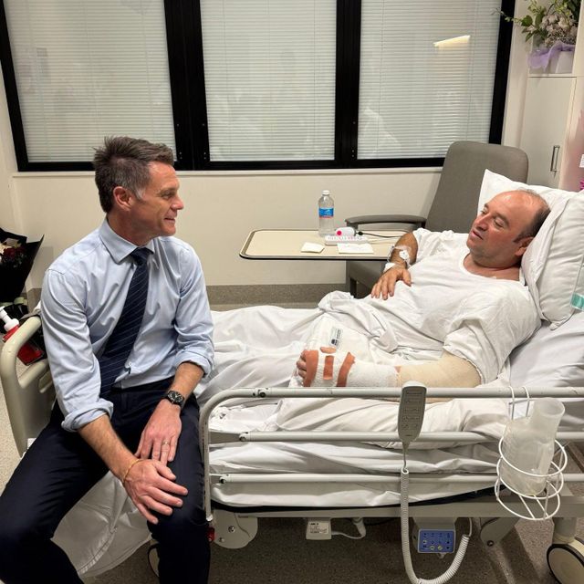 왼팔에 붕대를 감고 병상에 누워있는 아흐메드와 침대 끝에 앉아 있는 크리스 민스 뉴사우스웨일스 주지사