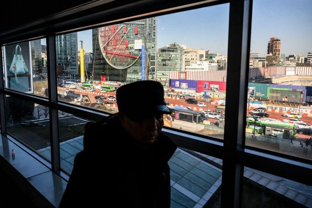 유리창 너머로 서울 도심의 번화한 거리와 고층 건물이 보인다. 창문 안쪽에는 모자를 쓴 한 남성이 서 있고, 바깥에는 차량과 버스가 도로를 따라 이동하고 있다. 