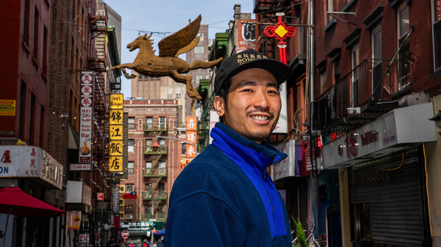 도심 거리에서 한 남성이 카메라를 향해 서 있다. 그 뒤로 중국어 간판과 장식물이 걸린 건물이 보인다.