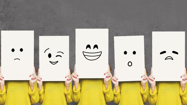 밝은 노란색 점퍼를 입은 다섯 명의 여성이 일렬로 서서 각기 다른 감정을 표현하는 커다란 직사각형의 흰색 종이를 얼굴에 뒤집어쓰고 있다