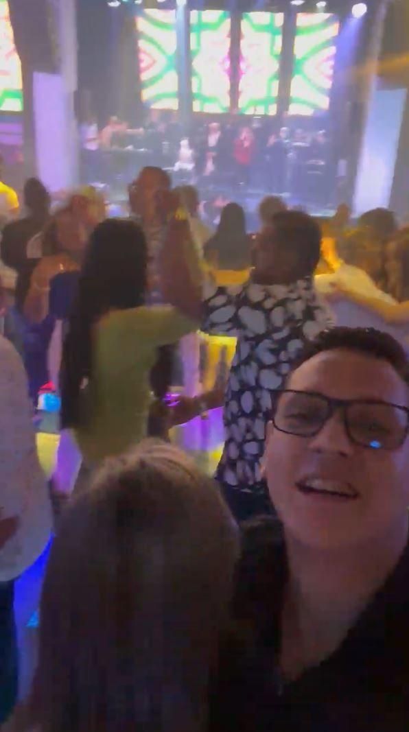 짧은 머리의 안경 쓴 남성이 사진을 찍고 있다. 배경에는 춤을 추고 있는 사람들과 멀리 위치한 무대.