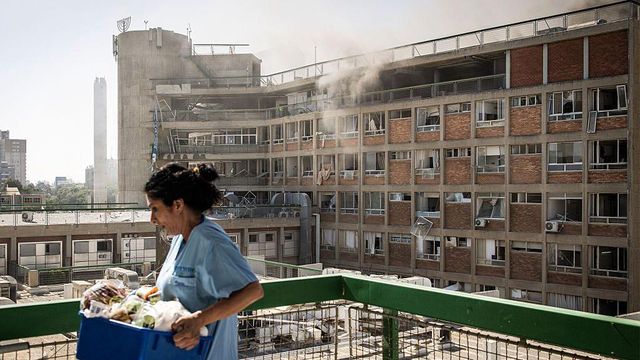 2025년 6월 19일 이스라엘 남부 베르셰바에 위치한 소로카 병원에서 이란의 미사일 공격 이후 연기가 피어오르는 건물 옆을 한 간호사가 의료 보급품을 들고 지나가고 있다