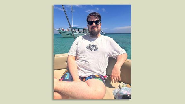 박쥐 그림이 그려진 흰색 배트맨 티셔츠와 공룡 무늬 반바지를 입은 남성이 바다 위 보트에 앉아 있다.