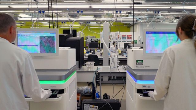 연구원 2명이 냉장고처럼 생긴 흰색의 고성능 시퀀싱 장비 앞에 등을 보이고 서 있다. 장비 위에는 컴퓨터 화면이 있다