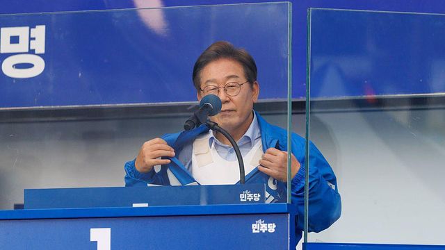 방탄유리 뒤 연단에 선 이재명 지사가 파란색 운동복 재킷을 열어 안에 입은 방탄조끼를 보여주고 있다