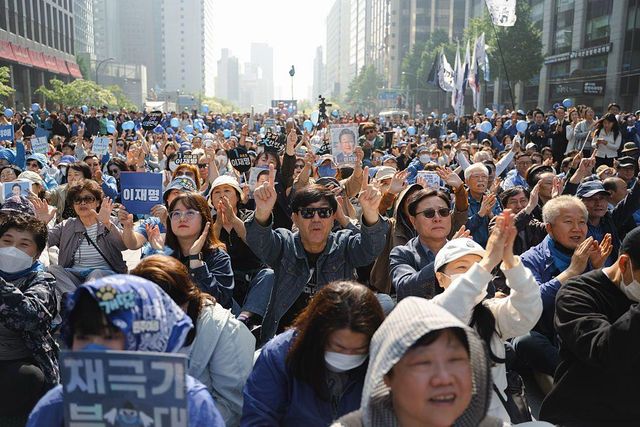 높고 현대적인 건물이 늘어선 시내 거리에 많은 이재명 지지자들이 모였고, 그중 일부는 파란색 플래카드를 들고 있다