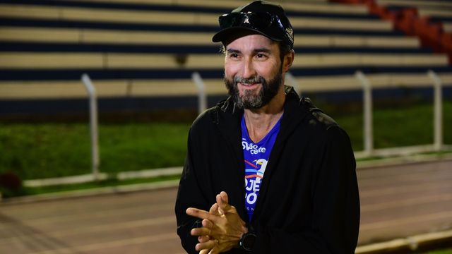 우고 파리아스는 파란색 운동복 상의에 검은색 재킷, 검은색 모자와 그 위에 선글라스를 썼다. 두 손을 모아 앞에 가지런히 놓고, 카메라 밖을 바라보며 미소 짓고 있다. 그는 육상 트랙 앞에 서 있다.