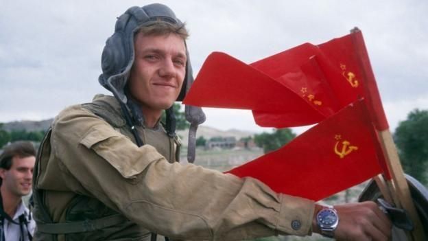 아프가니스탄에서 젊은 소련 군인이 웃으며 소련 국기 앞에서 포즈를 취하고 있다