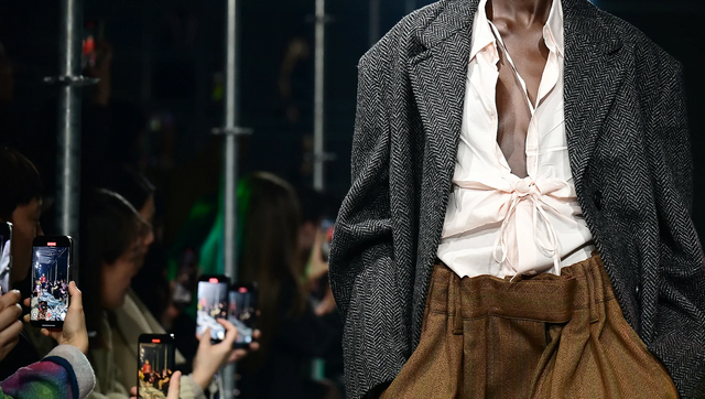 패션쇼 중 옆에는 관람객들이 휴대전화를 들고 영상을 찍고 있고, 한 모델이 런웨이를 걷고 있다. 