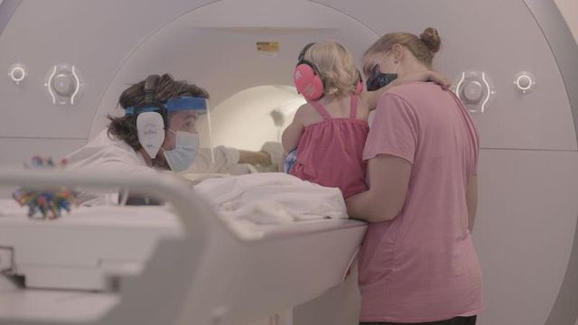 귀마개를 착용한 아기가 기능성 MRI 기기 옆에서 어머니에게 안겨 있고, 맞은편에는 연구자 터크-브라운 교수가 앉아 있다