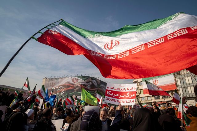 이란 시위 현장에서 대형 국기가 하늘에 펄럭이고, 아래에는 이란 국기를 든 시민들이 모여 있다. 배경에는 건물 외벽에 걸린 대형 현수막이 보인다