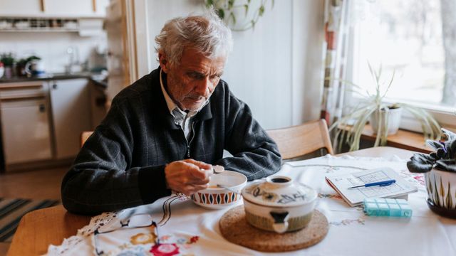 회색 머리의 노인이 집 안 식탁에 앉아 그릇에 담긴 시리얼을 먹고 있다. 