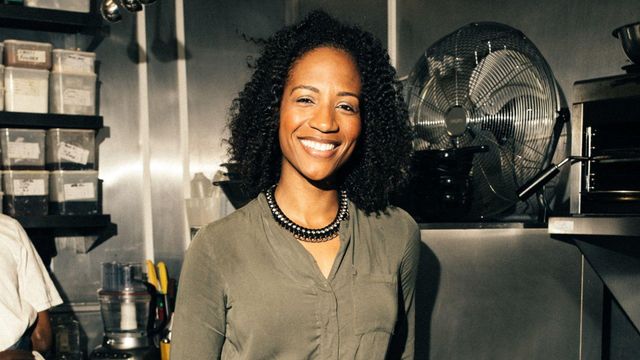 검은 곱슬머리를 한 한 여성이 카메라를 향해 미소를 짓고 있다. 그는 부엌에서 큰 검은색 선풍기 옆에 서 있다. 뒤편에는 각종 주방 도구들이 놓여 있으며, 그 뒤에서 작업 중인 것으로 보이는 남성의 팔꿈치가 살짝 보인다.