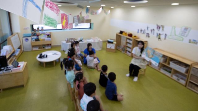 유치원 교실에서 한 교사가 십여명의 아이들 앞에 앉아서 큰 그림 책을 한쪽 팔에 들고 읽어주고 있다 (이 사진은 해당 사건과 관련이 없습니다)