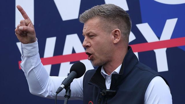 조끼를 입은 한 남성이 마이크를 들고 청중에게 연설하고 있다