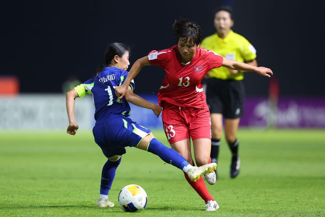 두 선수가 공을 두고 몸싸움을 벌이고 있다. 파란색 유니폼을 입은 한국 선수가 태클을 시도하고 있고, 빨간색 유니폼을 입은 북한 선수가 공을 지키려 하고 있다. 뒤편에는 심판이 경기를 지켜보고 있다. 