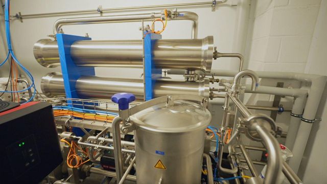 여러 개의 은색 탱크와 원통형 장치들이 배관으로 서로 연결된 채 흰 벽돌로 된 공간 안에 서 있으며, 왼쪽에는 전선과 기계 장비가 보인다