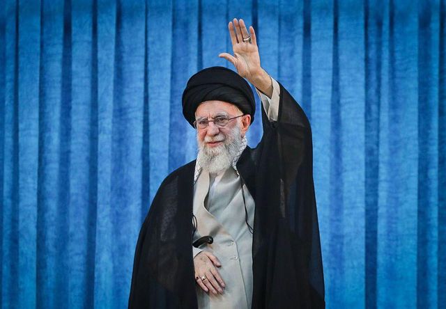 2025년 6월 4일 행사에서 전 이란 최고지도자 아야톨라 알리 하메네이가 파란색 커튼 앞에 서서 왼손을 들어 올리고 있는 모습