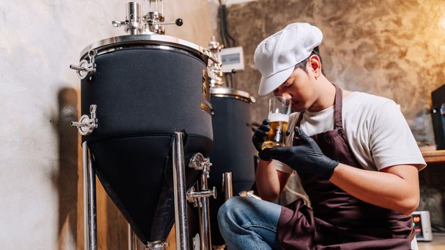 한 젊은 남성이 수제 맥주 양조장에서 작은 검은색 탱크 옆에 무릎을 꿇고 앉아, 잔에 담긴 맥주 샘플의 향을 맡고 있다
