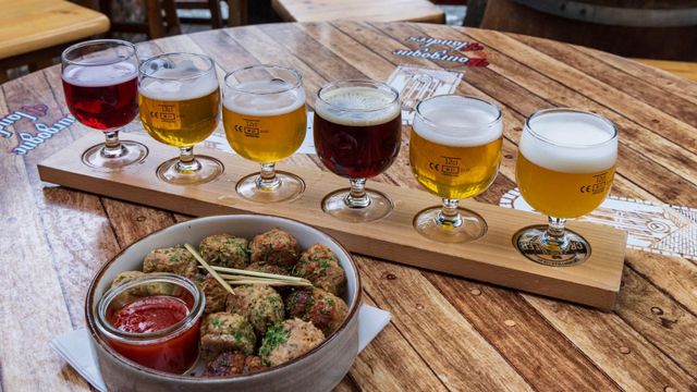 원형 나무 테이블 위에 놓인 나무 플래터에는 붉은색부터 황금빛까지 다양한 색의 맥주가 담긴 작은 잔 여섯 개가 있고 앞쪽에는 미트볼과 소스가 담긴 둥근 접시가 놓여 있다