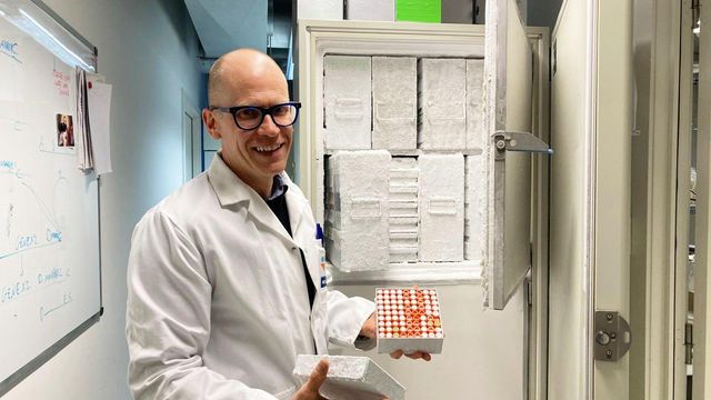 흰 실험복과 검은 안경을 쓴 한 남성이 수많은 용기로 가득 찬 큰 개방형 냉동고 앞에 서서 카메라를 향해 미소 짓고 있으며 서리가 덮인 금속 상자를 들고 있는데 그 안에는 작은 바이알들이 많이 들어 있다