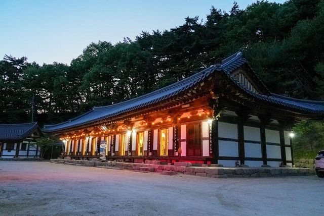 해 질 무렵 따뜻한 조명에 비친 전통 한국 사찰. 숲으로 우거진 산비탈 가장자리의 조용한 마당에 자리하고 있다. 