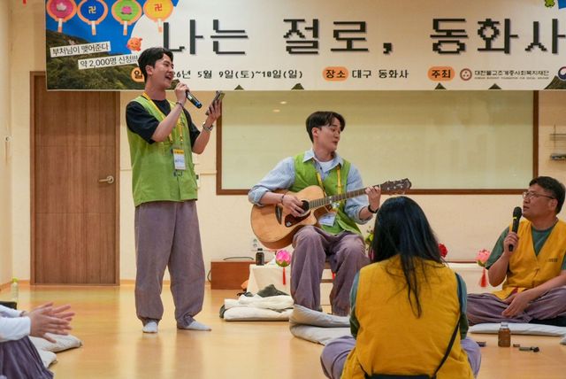 참가자들이 사찰 소개팅 수련회에서 음악을 연주하고 있다. 한 사람은 마이크를 들고 서 있고, 다른 사람은 앉아서 어쿠스틱 기타를 연주한다. 나머지 참가자들은 원형으로 놓인 방석에 앉아 있다. 뒤쪽에는 대형 현수막이 걸려 있다.
