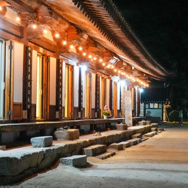 밤이 되자 따뜻한 조명이 켜진 한국의 전통 사찰 건물. 기와지붕과 나무 기둥, 넓은 마당을 따라 놓인 돌계단이 보이며 방문객들이 사찰 옆길을 걷고 있다