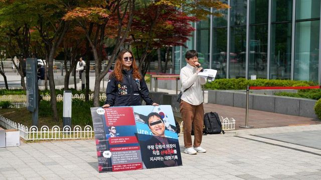 두 여성이 건물 앞에서 기자회견을 열고 있다. 한 명은 마이크를 잡고 발언하고, 다른 한 명은 손팻말을 들고 시위에 참여하고 있다.