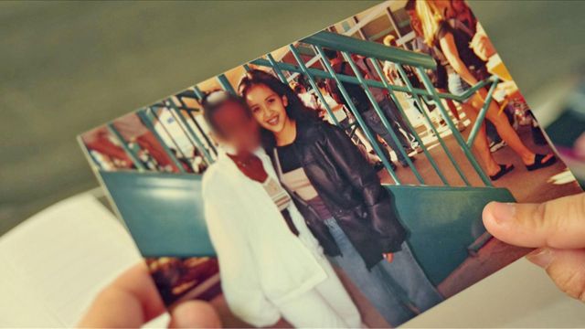 두 손으로 들고 있는 인쇄 사진. 사진에는 십 대 시절의 이자벨이 얼굴이 흐리게 처리된 친구와 함께 웃으며 포즈를 취하고 있다. 두 사람은 사람들이 붐비는 공공장소의 짙은 파란색 금속 난간 근처에 있다. 이자벨은 어두운색 재킷과 청바지를 입고 있으며, 얼굴이 흐려진 인물은 흰색 옷을 입고 있다.