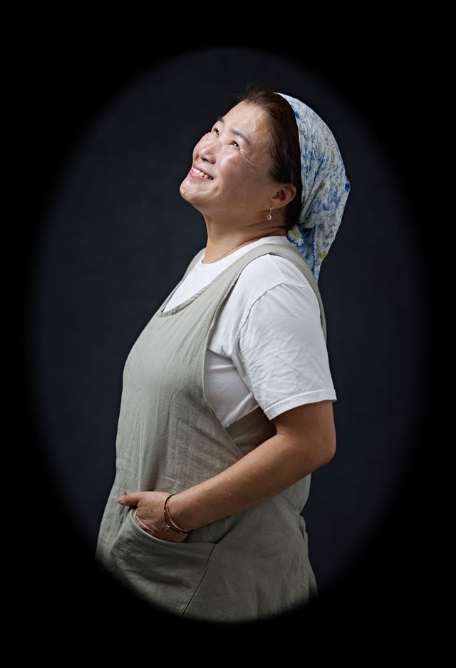 서대호 사진작가가 포착한 이정아(56)씨의 초상. 머릿수건을 두르고 앞치마 주머니에 손을 얹은 채 위를 바라보며 옅은 미소를 짓고 있다. 생계와 아이들의 돌봄으로 분주했던 일상의 차림 위로 고단한 날들 속에서도 웃음을 잃지 않았던 그의 온기가 묻어난다. ⓒ서대호 작가<br>