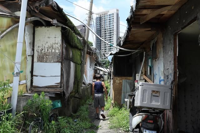 Guryong Village resident Cheon Yeong-hwan walks through a narrow alley between aging shacks. High-rise apartments loom beyond the alley, highlighting the contrast between Guryong Village and its surrounding neighborhoods. AJP Han Jun-gu