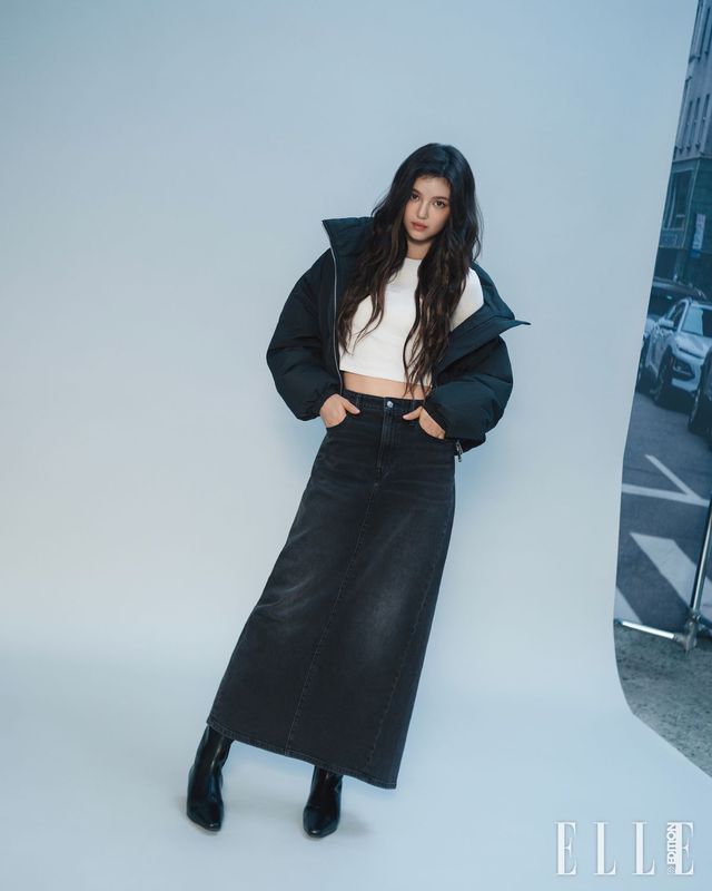 다니엘이 입은 화이트 롱 슬리브 모노그램 톱, 크롭 다운 푸퍼 재킷, 맥시 길이의 데님 스커트는모두 Calvin Klein Jeans.