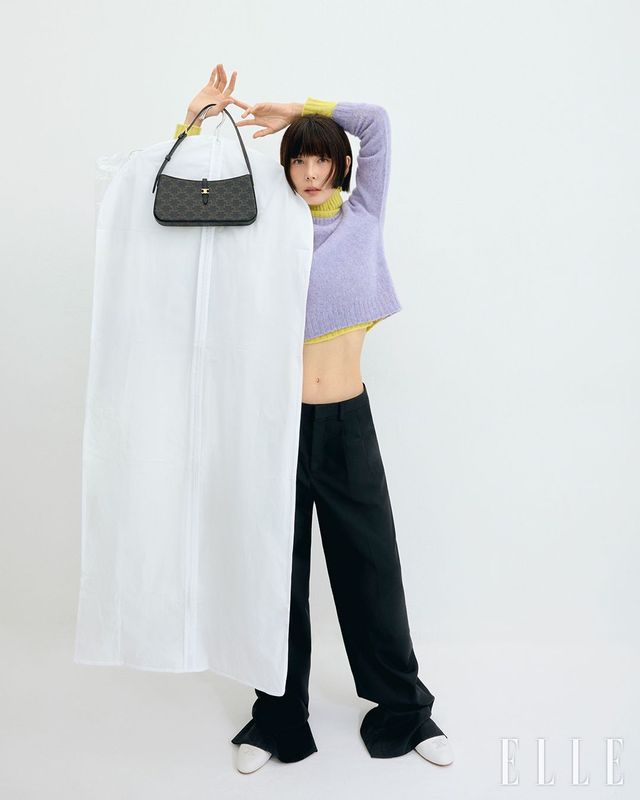 트리옹프 캔버스 티나 숄더백, 옐로 컬러의 셰틀랜드 울 터틀넥 스웨터와 레이어드 한 라벤더 컬러 울 크루넥 스웨터, 플리츠 트라우저, 트리옹프 화이트 램스킨 슈즈는 모두 Celine.