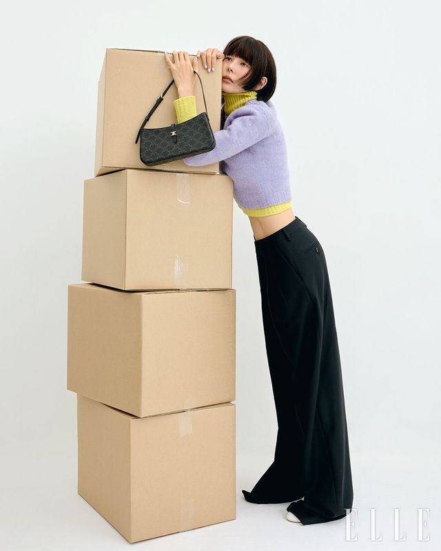 트리옹프 캔버스 티나 숄더백, 옐로 컬러의 셰틀랜드 울 터틀넥 스웨터와 레이어드한 라벤더 컬러 울 크루넥 스웨터, 플리츠 트라우저, 트리옹프 화이트 램스킨 슈즈는 모두 Celine.