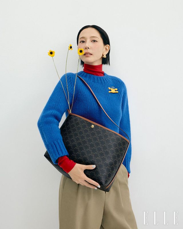 트리옹프 캔버스 & 카프스킨 소프트 쇼퍼백, 레드 컬러의 울 저지 터틀넥 톱과 레이어드한 캐시미어 실크 크루넥 스웨터, 코튼 울 소재의 플리츠 트라우저, 화이트 트리옹프 램스킨 슈즈, 폼 압스트레트 리본 후프 이어링은 모두 Celine.